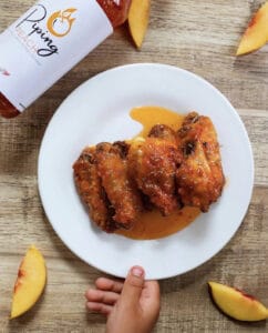 Ali di pollo su un piatto bianco seduto su un tavolo di legno circondato da fette di pesca e una bottiglia di salsa piccante Wing Suite Piping Peach