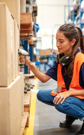 Warehouse worker crouching down and checking boxes on shelves with scanner. Female worker scanning boxes in rack.